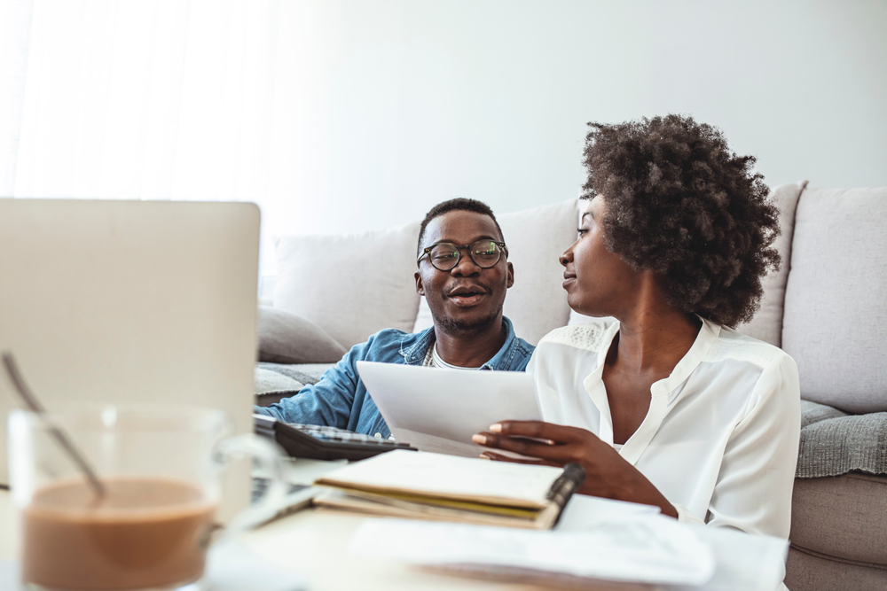 A young couple look at paperwork while sitting in front of a computer and doing their taxes online