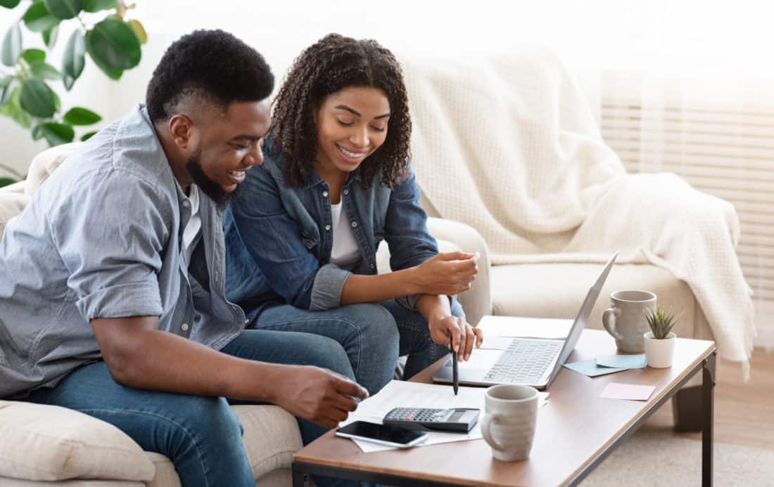 A man and woman sit on a couch while they look at a laptop computer that is sitting on the coffee table in front of them