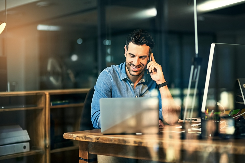 A man is shown taking on his mobile phone while looking at his computer. He's sitting at a wood table in a casual office setting.