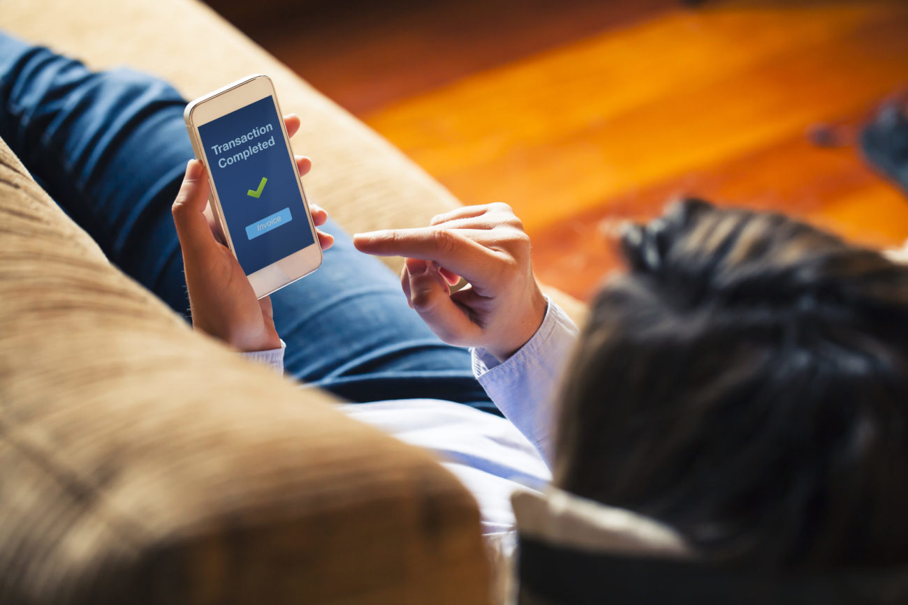 A woman reclining on a couch, viewed from above, uses her mobile phone for banking