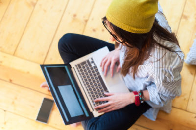 A young woman sits on the floor with her laptop computer and her smartphone as she engages in mobile banking