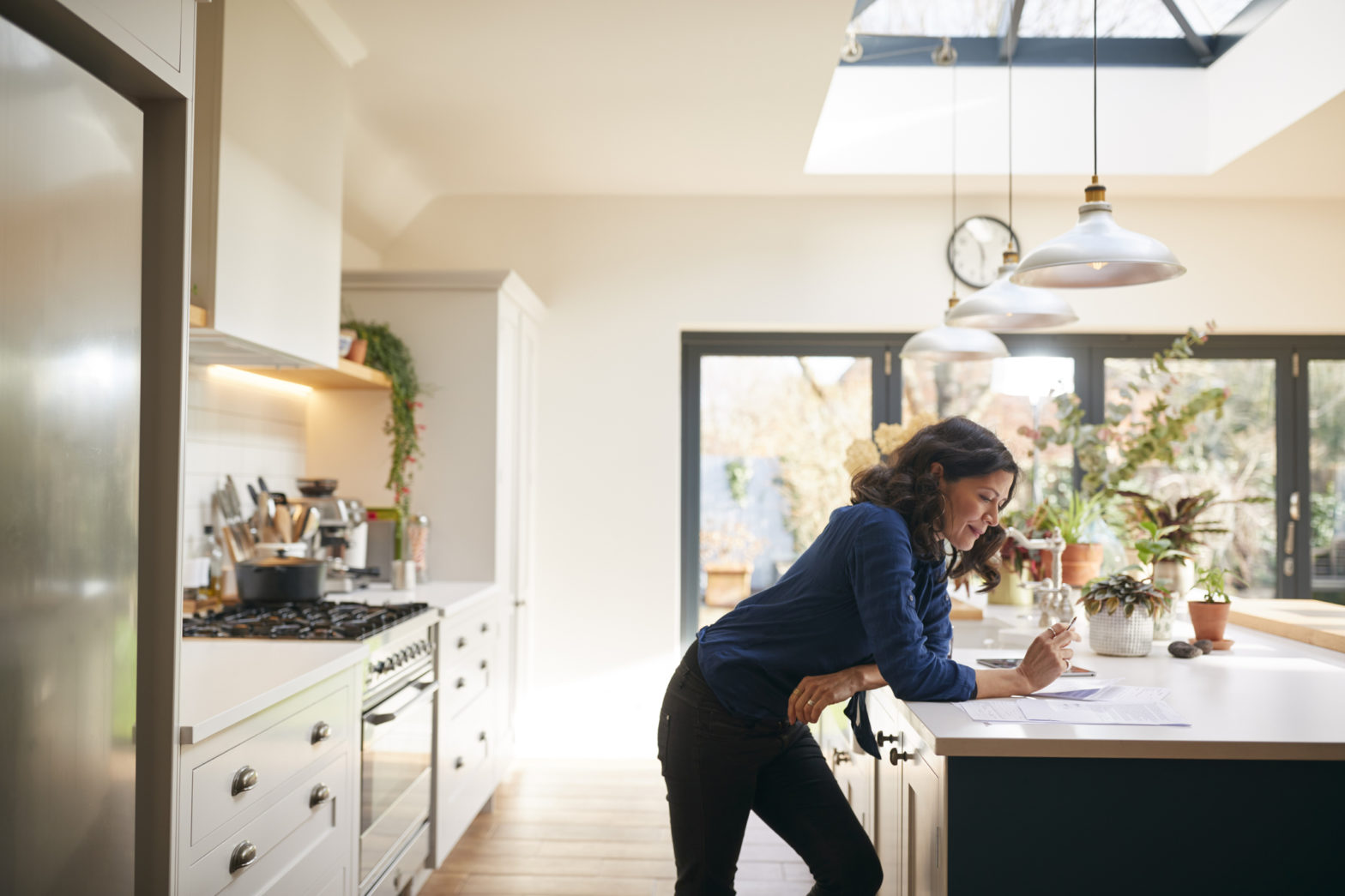 A woman leans on her kitchen counter as she looks at paperwork and works on her investments