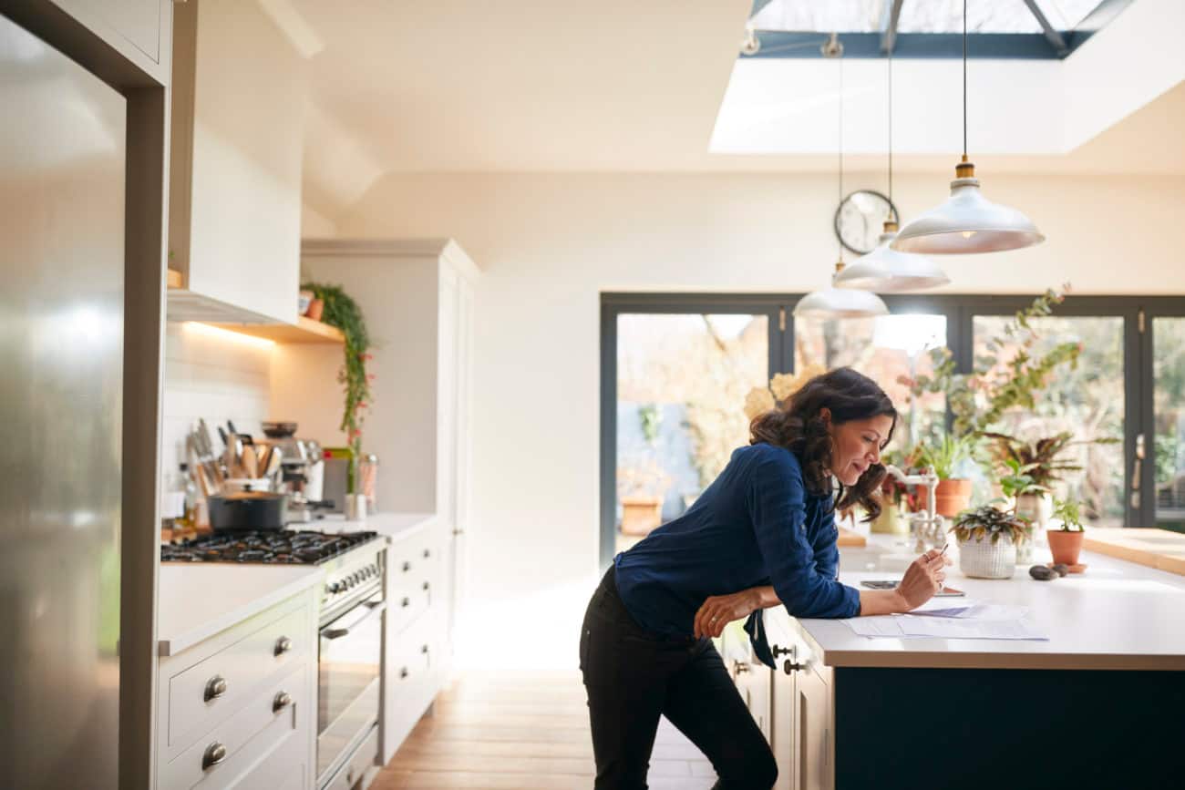 A woman leans on her kitchen counter as she looks at paperwork and works on her investments