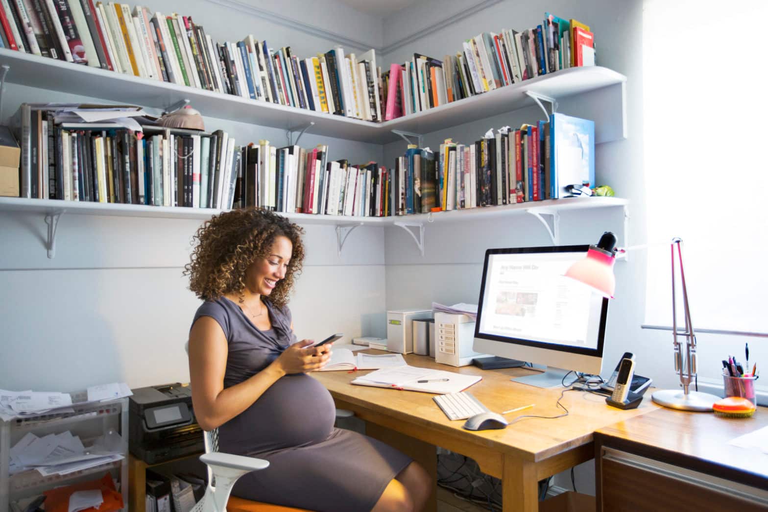 A pregnant woman sits in her home office and uses her cell phone to check on her savings account