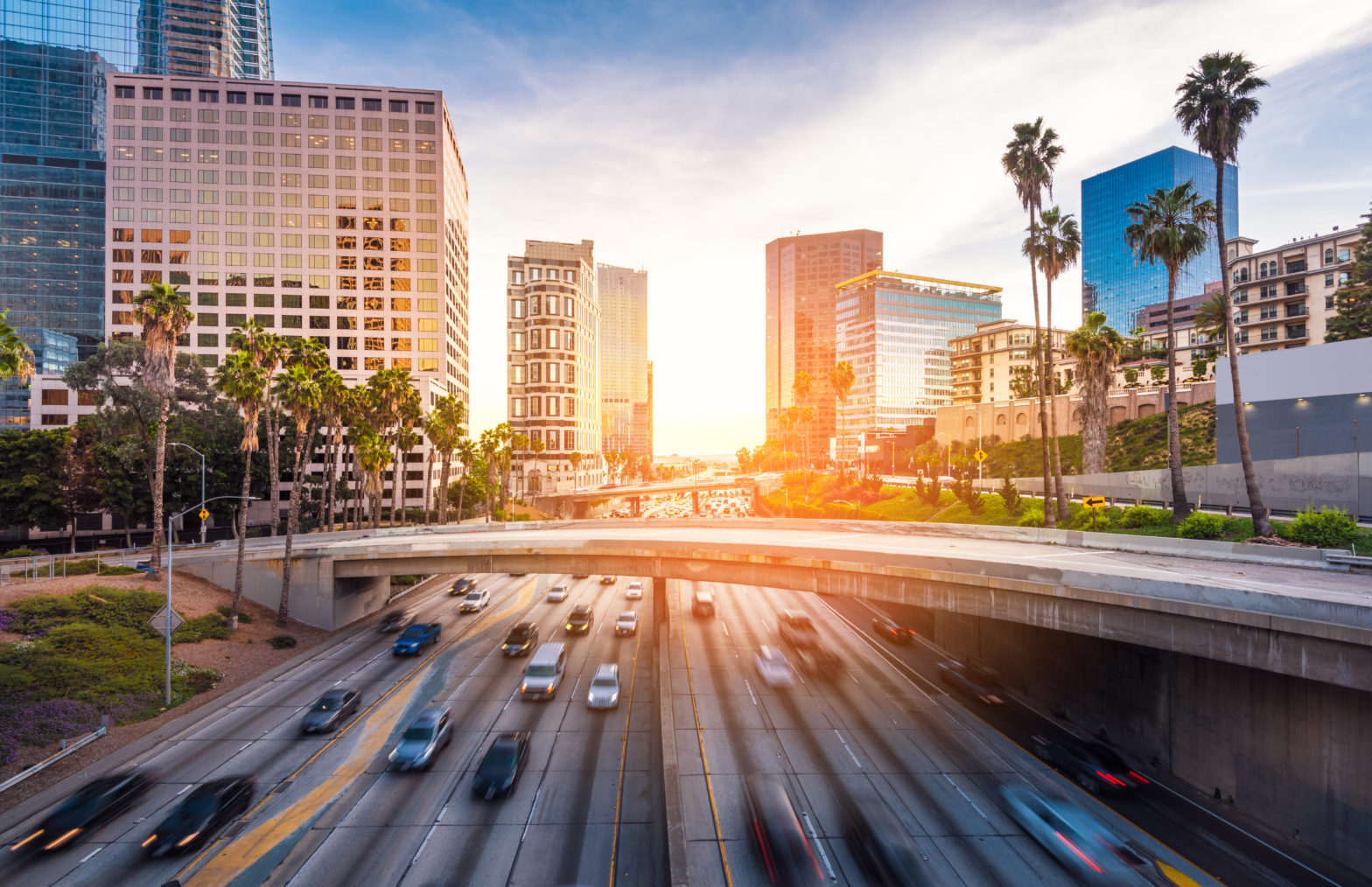 Sun setting between tall buildings while traffic whizzes by on the highway in Los Angeles.