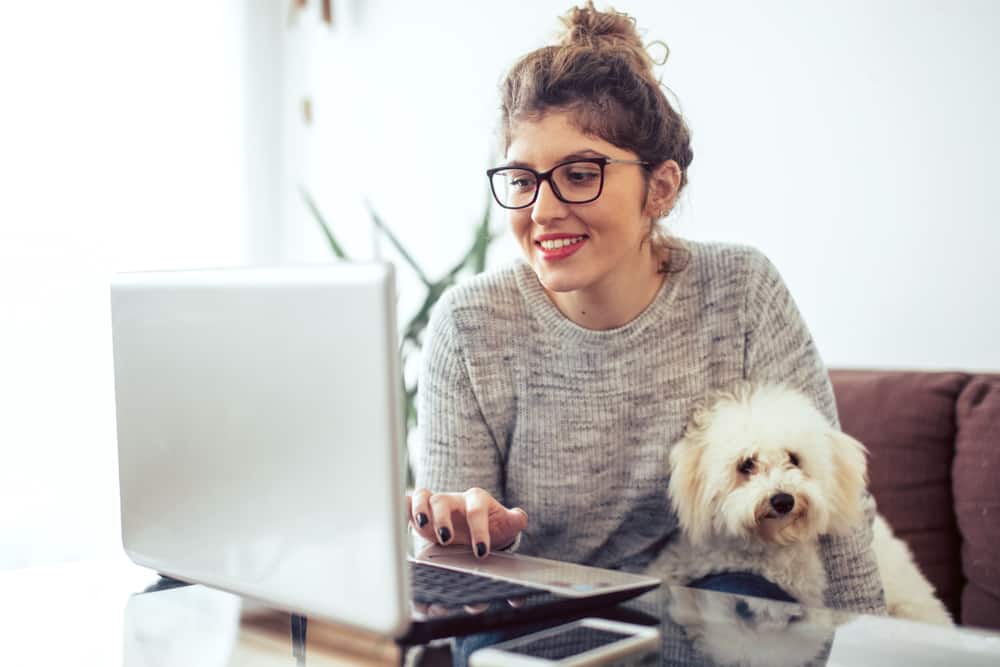 A young woman uses her laptop computer to bank from home while her dog sits next to her on the couch