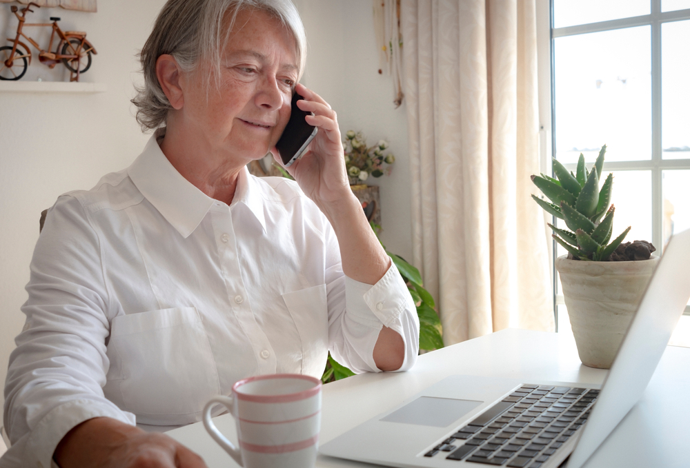 A woman sits at a desk in her home office looking at her computer and talking on the phone with her banker