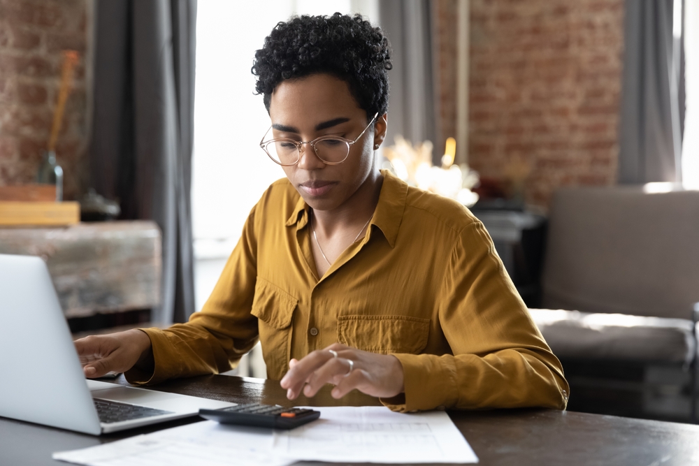 A young woman uses a calculator while looking at her savings account online to determine how much money she will earn in interest