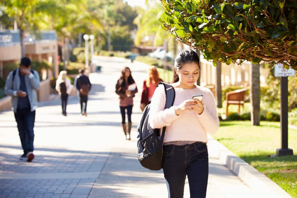 A woman with a backpack walks through a college campus looking at her savings account on her phone